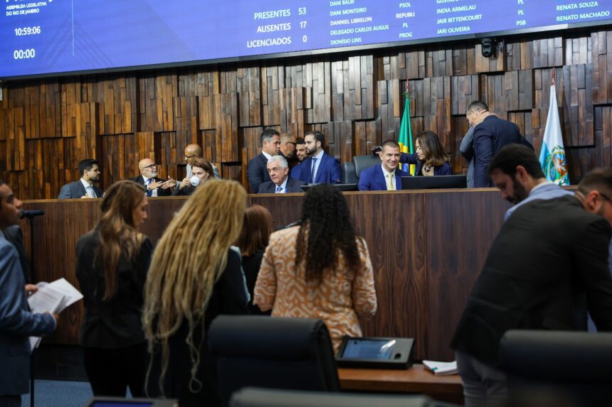 Plenário da Assembleia Legislativa do Estado do Rio de Janeiro durante sessão extraordinária. Parlamentares e assessores ocupam o plenário enquanto a Mesa Diretora conduz os trabalhos. Ao fundo, o painel eletrônico exibe o quórum da sessão e a presença dos deputados.