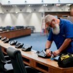 Funcionário realiza manutenção em um dos tablets instalados nas bancadas do plenário da Assembleia Legislativa do Estado do Rio de Janeiro (Alerj) durante o recesso parlamentar. Ao fundo, o plenário aparece desfocado enquanto outros trabalhadores atuam no local.