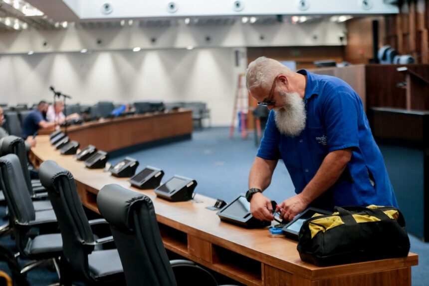 Funcionário realiza manutenção em um dos tablets instalados nas bancadas do plenário da Assembleia Legislativa do Estado do Rio de Janeiro (Alerj) durante o recesso parlamentar. Ao fundo, o plenário aparece desfocado enquanto outros trabalhadores atuam no local.