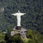 Vista aérea do Cristo Redentor no alto do Morro do Corcovado, cercado pela vegetação da Floresta da Tijuca. A plataforma de visitação está cheia de turistas ao redor do monumento.