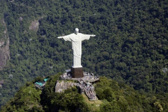 Vista aérea do Cristo Redentor no alto do Morro do Corcovado, cercado pela vegetação da Floresta da Tijuca. A plataforma de visitação está cheia de turistas ao redor do monumento.