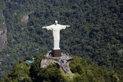 Vista aérea do Cristo Redentor no alto do Morro do Corcovado, cercado pela vegetação da Floresta da Tijuca. A plataforma de visitação está cheia de turistas ao redor do monumento.