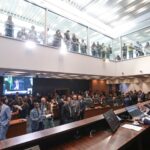 Vista ampla do plenário da Assembleia Legislativa do Estado do Rio de Janeiro (Alerj) durante sessão deliberativa. Deputados e assessores conversam no plenário, enquanto servidores acompanham a votação da galeria envidraçada segurando cartazes. Ao fundo, um telão exibe a transmissão da sessão, e o presidente da Mesa conduz os trabalhos.