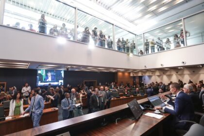 Vista ampla do plenário da Assembleia Legislativa do Estado do Rio de Janeiro (Alerj) durante sessão deliberativa. Deputados e assessores conversam no plenário, enquanto servidores acompanham a votação da galeria envidraçada segurando cartazes. Ao fundo, um telão exibe a transmissão da sessão, e o presidente da Mesa conduz os trabalhos.