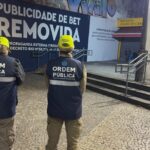 Dois agentes da Secretaria Municipal de Ordem Pública observam um painel onde a publicidade de uma plataforma de apostas foi coberta pela mensagem "Publicidade de Bet Removida", em área comercial do Rio de Janeiro, após a entrada em vigor do decreto que proíbe esse tipo de anúncio em espaços públicos.