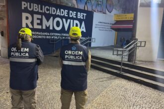 Dois agentes da Secretaria Municipal de Ordem Pública observam um painel onde a publicidade de uma plataforma de apostas foi coberta pela mensagem "Publicidade de Bet Removida", em área comercial do Rio de Janeiro, após a entrada em vigor do decreto que proíbe esse tipo de anúncio em espaços públicos.