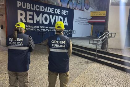 Dois agentes da Secretaria Municipal de Ordem Pública observam um painel onde a publicidade de uma plataforma de apostas foi coberta pela mensagem "Publicidade de Bet Removida", em área comercial do Rio de Janeiro, após a entrada em vigor do decreto que proíbe esse tipo de anúncio em espaços públicos.