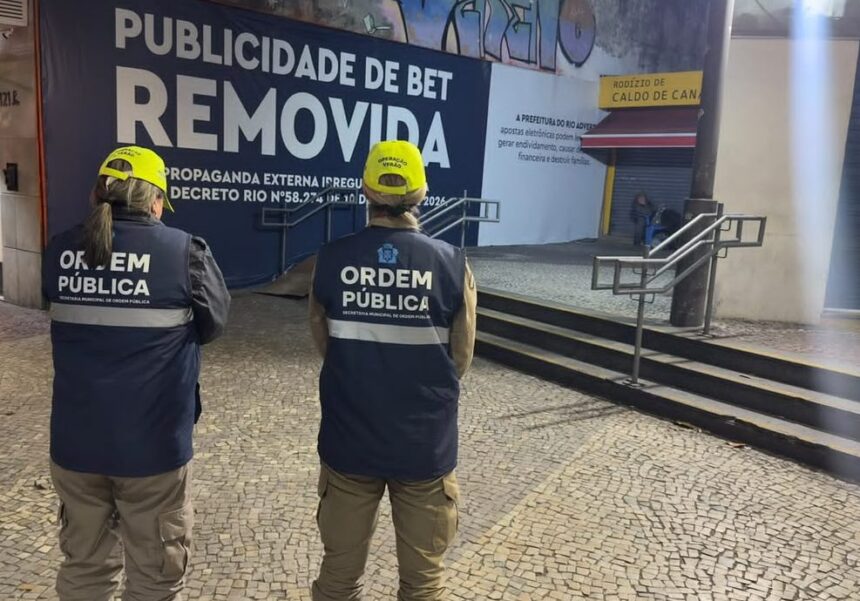 Dois agentes da Secretaria Municipal de Ordem Pública observam um painel onde a publicidade de uma plataforma de apostas foi coberta pela mensagem "Publicidade de Bet Removida", em área comercial do Rio de Janeiro, após a entrada em vigor do decreto que proíbe esse tipo de anúncio em espaços públicos.