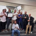 Eduardo Paes posa ao lado de vereadores e apoiadores durante cerimônia na Câmara Municipal de Seropédica, segurando uma moção de aplausos recebida em homenagem aos serviços prestados. Os participantes aparecem aplaudindo em um plenário, enquanto uma pessoa registra o momento com um celular.