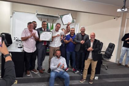 Eduardo Paes posa ao lado de vereadores e apoiadores durante cerimônia na Câmara Municipal de Seropédica, segurando uma moção de aplausos recebida em homenagem aos serviços prestados. Os participantes aparecem aplaudindo em um plenário, enquanto uma pessoa registra o momento com um celular.
