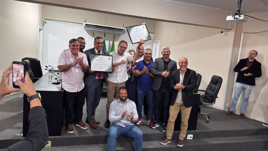 Eduardo Paes posa ao lado de vereadores e apoiadores durante cerimônia na Câmara Municipal de Seropédica, segurando uma moção de aplausos recebida em homenagem aos serviços prestados. Os participantes aparecem aplaudindo em um plenário, enquanto uma pessoa registra o momento com um celular.