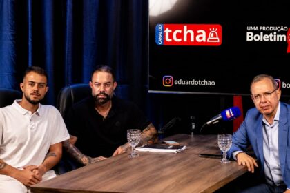 Poubel e o filho, pré-candidato à Alerj, posam ao lado do jornalista Eduardo Tchao durante a gravação do Boletim Cast, em estúdio com mesa, microfones e painel do programa ao fundo.