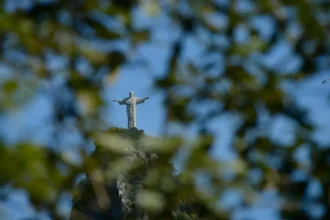Cristo Redentor visto entre folhas de árvores, no alto do Morro do Corcovado, sob o céu azul.