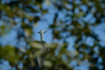 Cristo Redentor visto entre folhas de árvores, no alto do Morro do Corcovado, sob o céu azul.