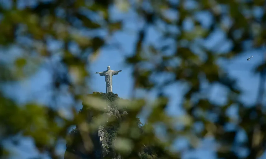 Cristo Redentor visto entre folhas de árvores, no alto do Morro do Corcovado, sob o céu azul.