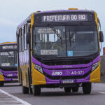 Ônibus novos da Prefeitura do Rio, nas cores roxa e amarela, circulam em fila por uma via de Santa Cruz.