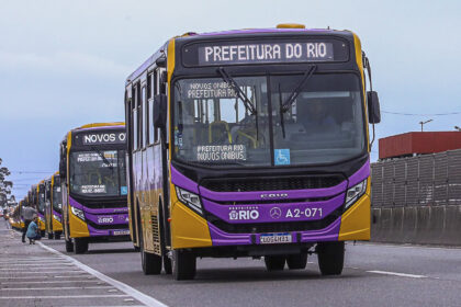 Ônibus novos da Prefeitura do Rio, nas cores roxa e amarela, circulam em fila por uma via de Santa Cruz.
