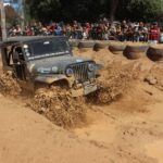 Jipe atravessa uma pista de lama durante o Encontro Nacional de Jipeiros em Atafona, acompanhado pelo público.