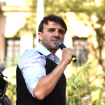 Renan Santos discursa ao microfone durante ato público ao ar livre, vestindo camisa de manga longa dobrada e colete escuro.