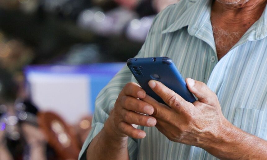 Close das mãos de um homem com camisa listrada segurando e manuseando um smartphone azul.
