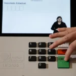 Mão pressiona o botão verde “Confirma” de uma urna eletrônica, que exibe uma intérprete de Libras na tela.