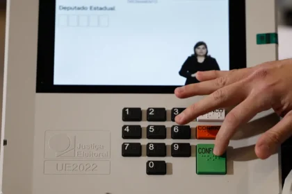 Mão pressiona o botão verde “Confirma” de uma urna eletrônica, que exibe uma intérprete de Libras na tela.