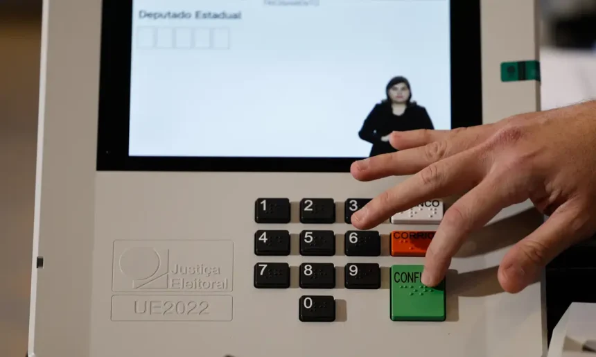 Mão pressiona o botão verde “Confirma” de uma urna eletrônica, que exibe uma intérprete de Libras na tela.