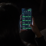 Mulher vista de costas usa um celular com uma plataforma de apostas esportivas aberta em um ambiente escuro.