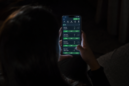 Mulher vista de costas usa um celular com uma plataforma de apostas esportivas aberta em um ambiente escuro.