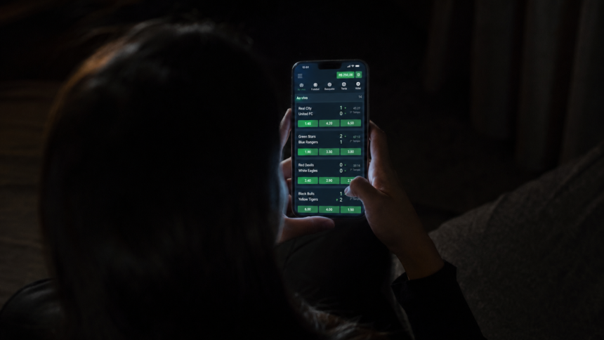 Mulher vista de costas usa um celular com uma plataforma de apostas esportivas aberta em um ambiente escuro.