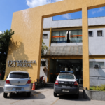 Fachada da 123ª Delegacia de Polícia de Macaé, com carros e uma viatura estacionados em frente ao prédio.