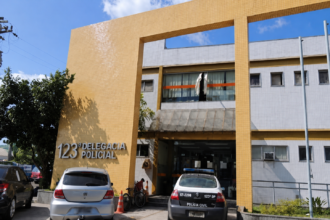 Fachada da 123ª Delegacia de Polícia de Macaé, com carros e uma viatura estacionados em frente ao prédio.