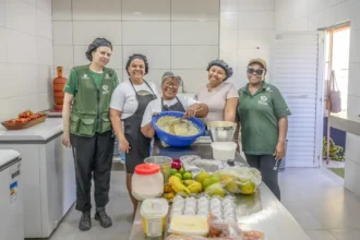 Cinco integrantes da Cozinha das Caranguejeiras posam em uma cozinha, ao redor de uma mesa com ingredientes e utensílios.