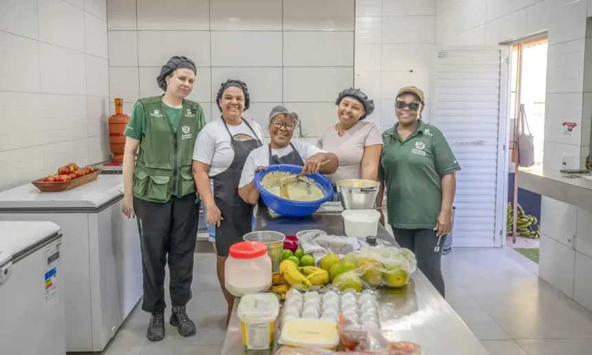 Cinco integrantes da Cozinha das Caranguejeiras posam em uma cozinha, ao redor de uma mesa com ingredientes e utensílios.