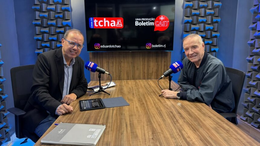 O jornalista Eduardo Tchao entrevista o empresário Eike Batista no estúdio do Boletim Cast. Os dois estão sentados frente a frente, diante de uma mesa com microfones, enquanto ao fundo um monitor exibe as marcas do Canal do Tchao e do Boletim Cast.