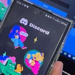 Celular exibe a tela inicial do Discord diante de um computador aberto na página de acesso à plataforma.