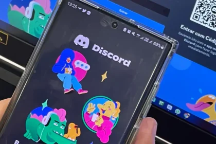 Celular exibe a tela inicial do Discord diante de um computador aberto na página de acesso à plataforma.
