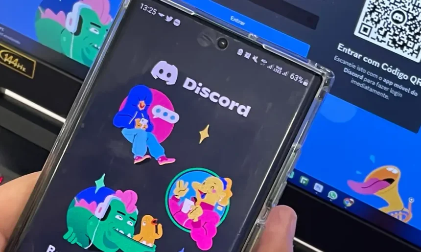 Celular exibe a tela inicial do Discord diante de um computador aberto na página de acesso à plataforma.