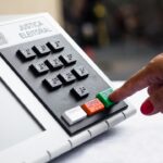 Mão com unhas pintadas de rosa pressiona a tecla verde “Confirma” em uma urna eletrônica.