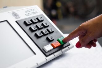 Mão com unhas pintadas de rosa pressiona a tecla verde “Confirma” em uma urna eletrônica.