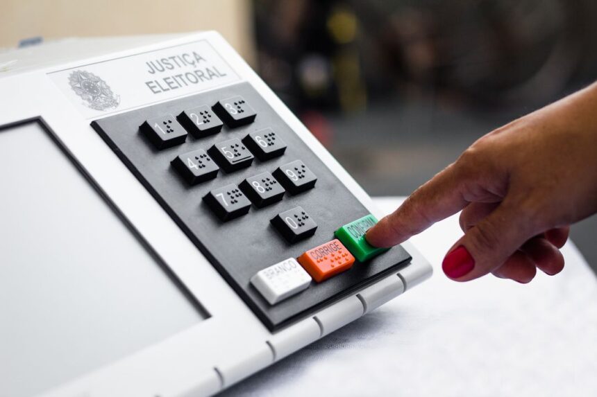 Mão com unhas pintadas de rosa pressiona a tecla verde “Confirma” em uma urna eletrônica.