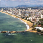 Vista aérea da orla e da área urbana de Macaé, com prédios, praias, mar e montanhas ao fundo.
