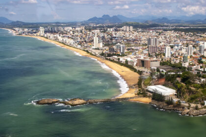 Vista aérea da orla e da área urbana de Macaé, com prédios, praias, mar e montanhas ao fundo.