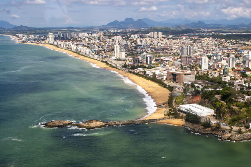 Vista aérea da orla e da área urbana de Macaé, com prédios, praias, mar e montanhas ao fundo.