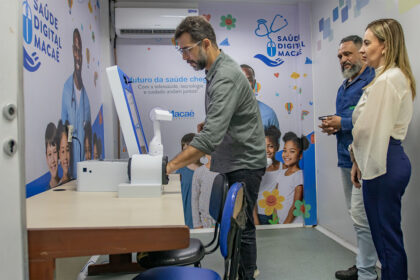Profissional demonstra equipamentos de Telessaúde em sala de atendimento pediátrico da UPA da Barra, em Macaé, enquanto outras duas pessoas observam.