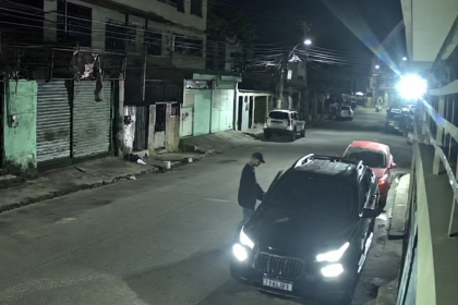 Imagem de câmera de segurança mostra uma pessoa ao lado de um veículo preto estacionado durante a noite.