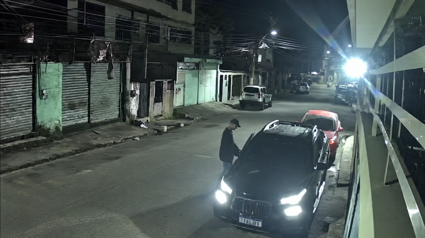 Imagem de câmera de segurança mostra uma pessoa ao lado de um veículo preto estacionado durante a noite.