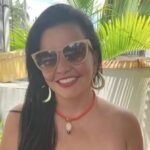 Karen Mancini sorri usando óculos escuros, brincos grandes e um colar vermelho, com plantas ao fundo.