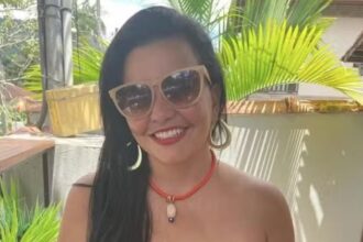 Karen Mancini sorri usando óculos escuros, brincos grandes e um colar vermelho, com plantas ao fundo.