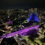 Vista aérea noturna dos Arcos da Lapa iluminados em tons de rosa e roxo, com a Catedral Metropolitana ao fundo.